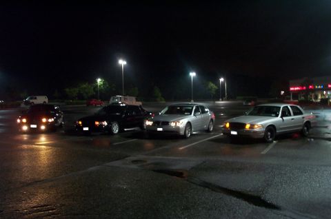1999 Ford Crown Victoria - Nighttime meet in a wet strip-mall parking lot with four sedans parked in a row, headlights on. A silver Ford Crown Victoria sits at right…