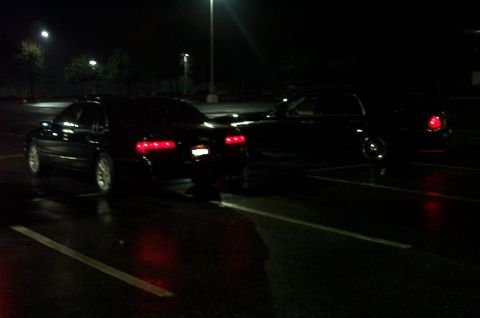 1999 Ford Crown Victoria - Night shot in a wet parking lot of a dark full-size sedan seen from the rear three-quarter with its tail lights and license plate lamp lit,…
