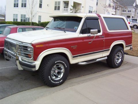 1986 Ford Bronco - This two-tone Ford Bronco is parked in front of a building with another car visible.