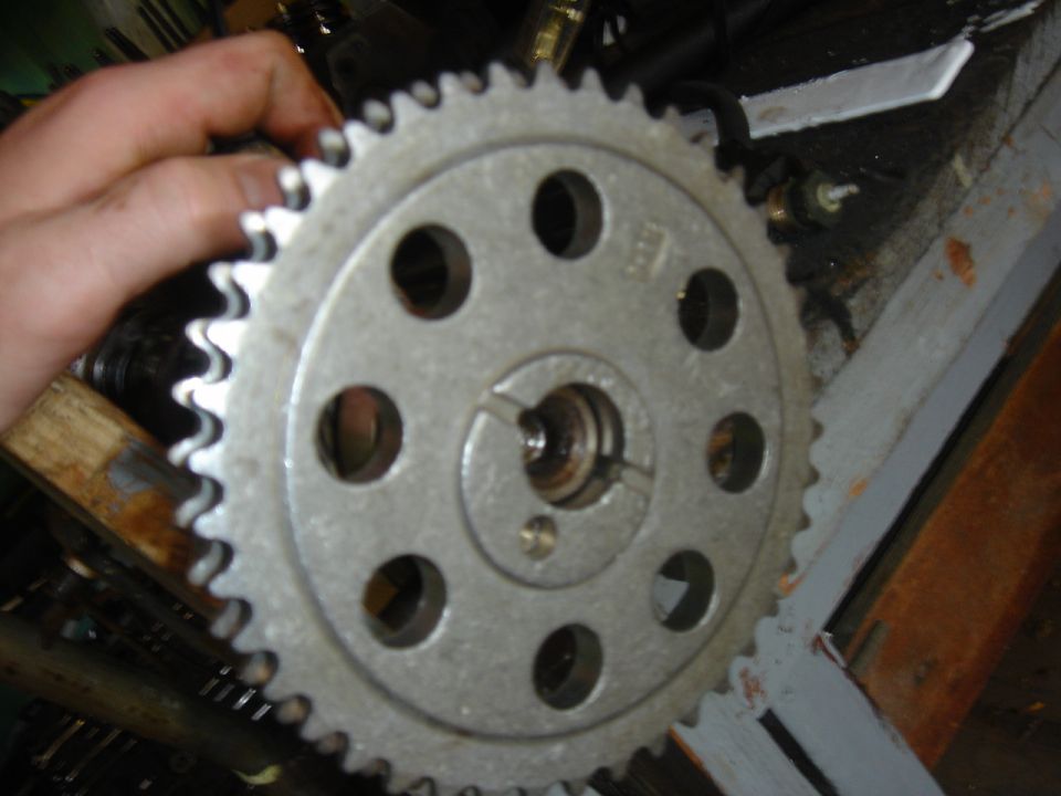 Close-up of a metal camshaft gear with teeth and bolt holes, held by a hand