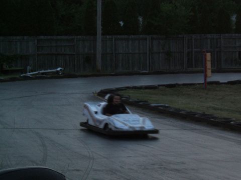 1998 Ford Taurus - A go-kart navigates a turn on a track, with a wooden fence and trees visible behind it.
