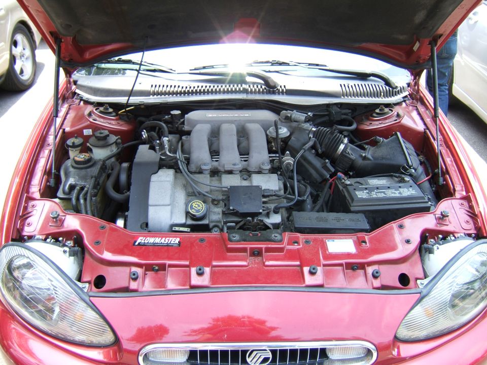 red car's engine bay with V6 engine, battery, and hoses visible. Another car is partially seen to the left