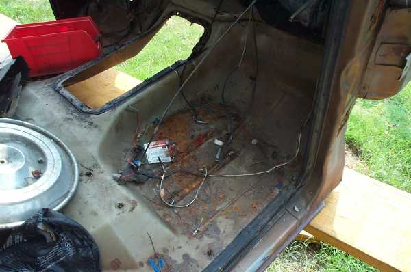 Disassembled vehicle interior showing rusted floor, wiring, and a red toolbox. Hubcap visible on left