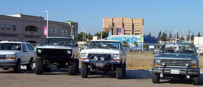 1995 Ford Bronco - A lineup of four lifted trucks, including a white Ford F-Series, are parked outdoors under a clear blue sky.