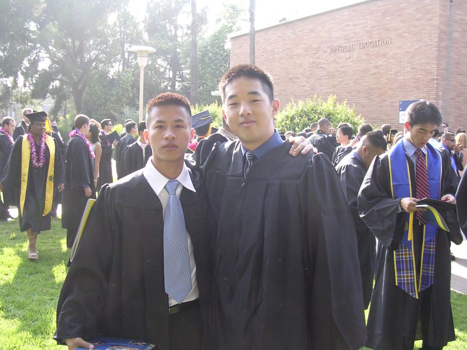 Two graduates in black gowns and blue ties pose for a photo on a sunny day, with other graduates in the background