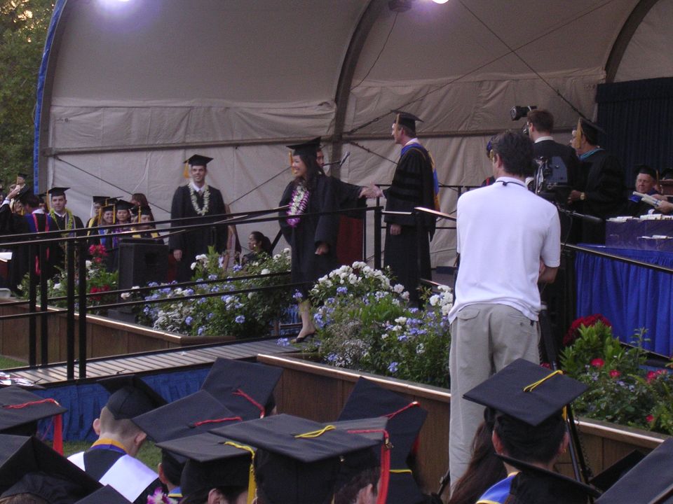 People in graduation gowns on a stage, some receiving leis. Audience in foreground wearing graduation caps