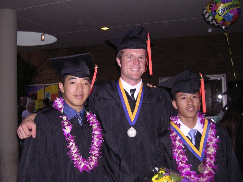Three young men in graduation gowns and leis pose for a photo with a 'Congratulations' balloon