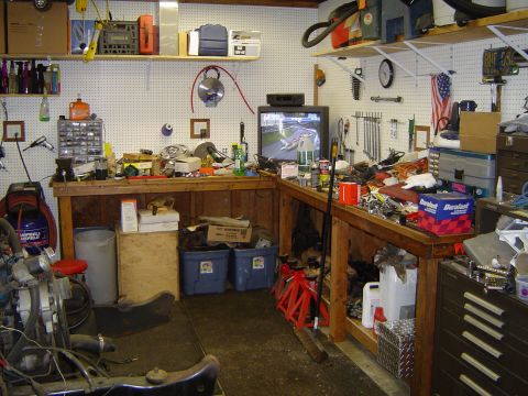 1989 Ford Bronco - A cluttered garage workbench is filled with tools, parts, and supplies, with a television set in the background showing a racing game.