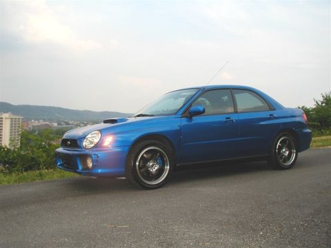 2002 Subaru Impreza - A blue 2002 Subaru Impreza sedan is captured from a 3/4 front angle, with a city visible in the distance.