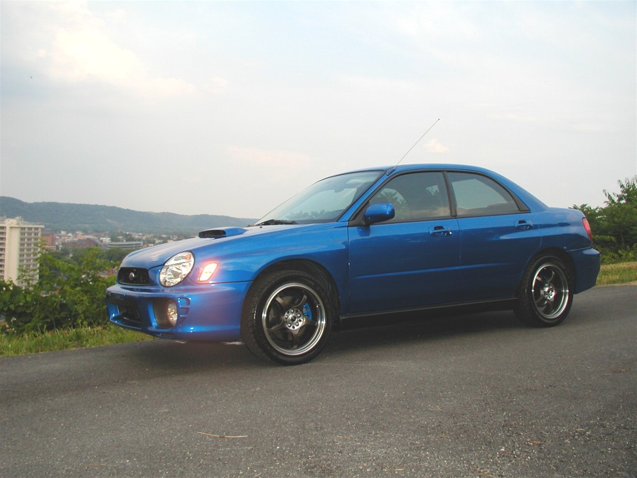 Blue 2002 Subaru Impreza sedan parked on asphalt with a city skyline in the background