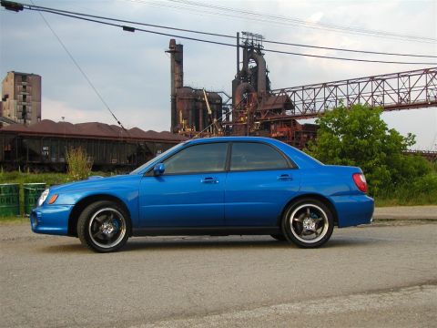 2002 Subaru Impreza - A blue 2002 Subaru Impreza sedan is parked in front of an industrial complex with train cars in the background.