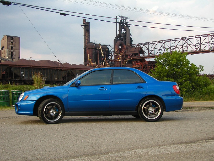 Blue Subaru Impreza sedan, side view, parked before an industrial steel mill backdrop.