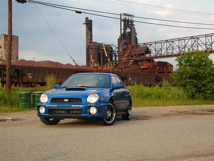 Blue Subaru Impreza with hood scoop parked before an old steel mill