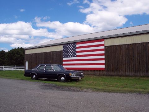 2001 Ford Crown Victoria - A dark sedan is parked on the grass next to a barn decorated with a large American flag mural.
