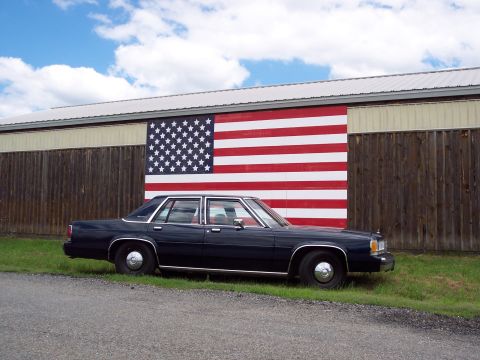 2001 Ford Crown Victoria - A dark blue sedan is parked on the grass in front of a building painted with a large American flag mural.