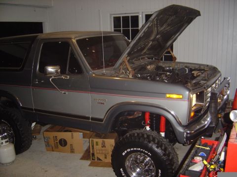 1986 Ford Bronco - A gray 1986 Ford Bronco is parked indoors with its hood open, revealing the engine bay and bright red shocks on the suspension.