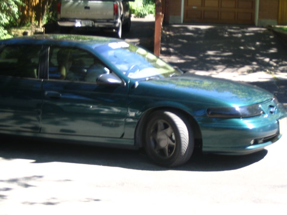 Side profile of teal-green Ford Taurus sedan in a driveway near wood-sided garage