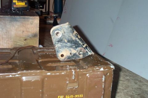 1995 Ford Bronco - Worn stamped-steel mounting bracket with a large bushing eye at the top and three lower bolt holes, its black paint flaking, sitting on a b…