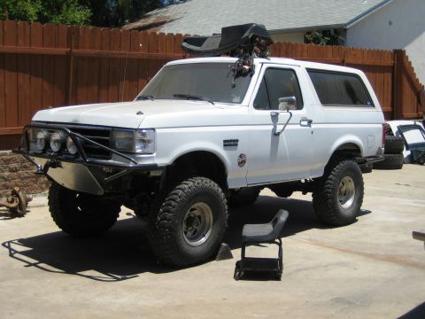1990 Ford Bronco - White lifted Ford Bronco on a driveway by a wood fence, with a tube front bumper, off-road lights and a tube seat nearby.