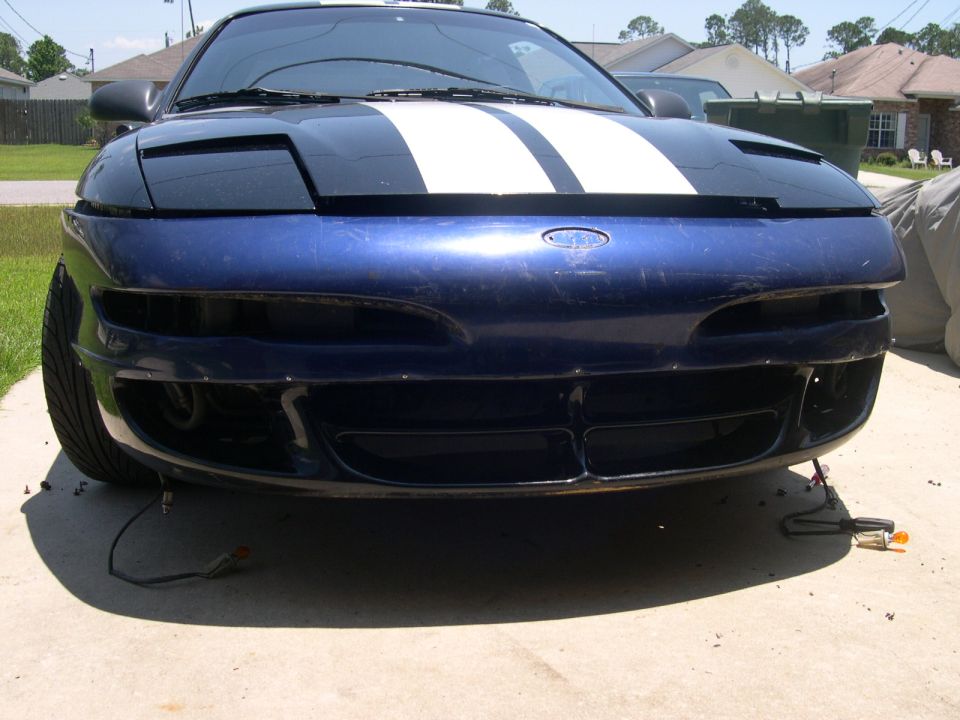 Front view of a blue car with white racing stripes on the hood. The bumper is partially removed