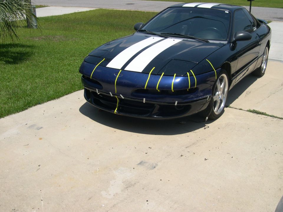 Blue 1997 Ford Probe with white racing stripes and yellow tape on the front bumper, parked on a driveway