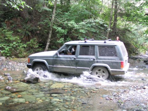 2000 Jeep Cherokee - A Jeep Cherokee navigates a stream in a forest, kicking up water as it goes.