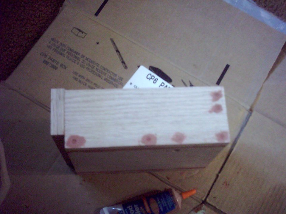 Unfinished wooden box with wood filler spots drying, on cardboard next to a filler bottle