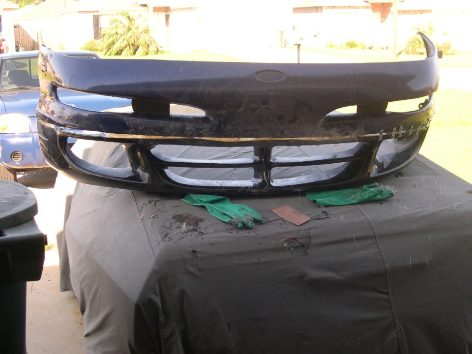 Dark colored car bumper with grille and headlight openings, resting on a covered surface. A blue vehicle is visible in the background