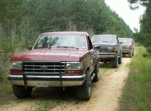 1988 Ford Bronco - Three Ford trucks, including a red 1988 Bronco, drive in a line on a dirt road through a wooded area.