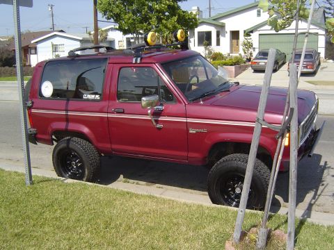 1985 Ford Bronco II - A red 1985 Ford Bronco II is parked on the side of the road, a roof rack and off-road tires.