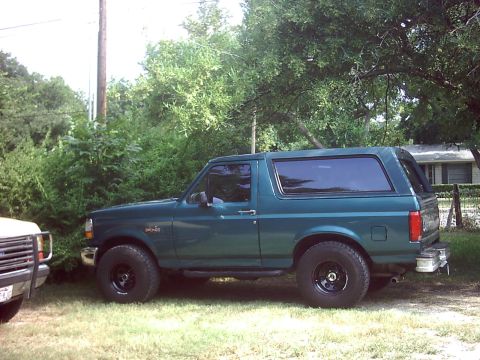 1996 Ford Bronco - Teal-green Ford Bronco shown in full driver-side profile on a grassy yard with trees overhead, riding on black steel wheels with all-terrai…