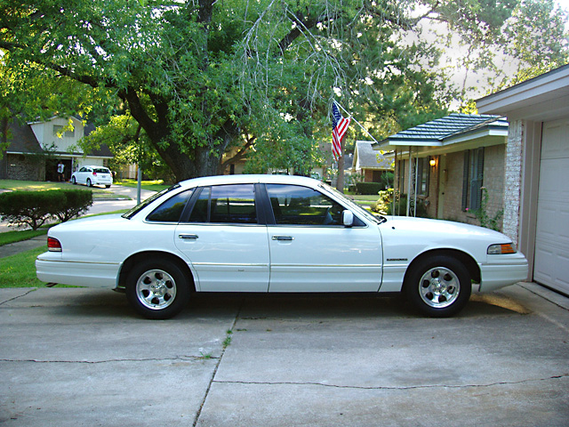 White Ford Crown Victoria sedan in side profile on a driveway with a house and American flag behind it