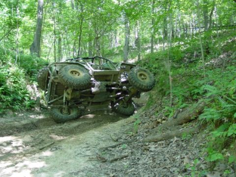 1992 Ford Bronco - An off-road vehicle has tipped onto its side while navigating a muddy path through a dense forest.