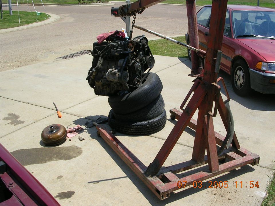Engine suspended from red hoist over stacked tires in driveway, torque converter on ground, red car nearby