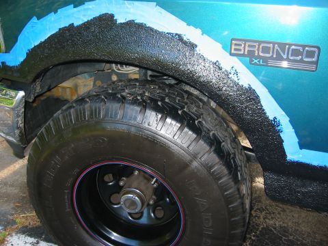 1996 Ford Bronco - Close-up of a teal Ford Bronco XL rear wheel arch mid-project, with black textured bed-liner coating being applied over blue painter's-tape…