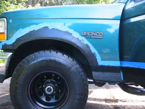 1996 Ford Bronco - Close-up of a teal/green Ford Bronco's front fender and wheel arch mid-restoration, with black textured bedliner (Herculiner) coating appli…
