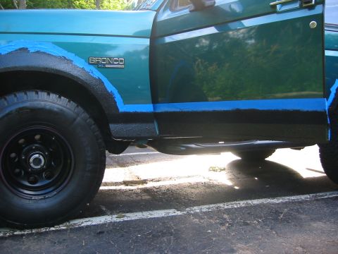 1996 Ford Bronco - Side view of a teal-green Ford Bronco XL's lower body mid-project, with blue masking tape marking a freshly rolled black bedliner coating a…
