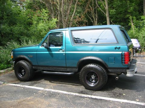 1996 Ford Bronco - Teal Ford Bronco with removable hardtop shown in profile from the driver's side in a wooded parking lot, wearing fresh black textured bedli…