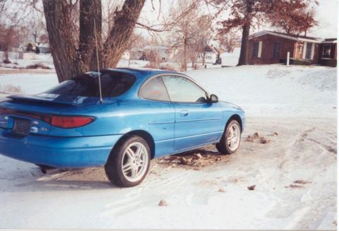 2000 Ford Escort - A blue coupe sits on a snow-covered ground with a house and trees in the background.