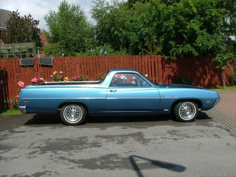 1970 Ford Ranchero - Side profile of a blue 1970 Ford Ranchero with a wooden fence and trees in the background.
