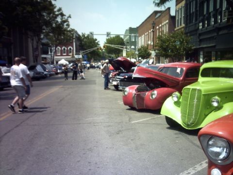 1995 Ford Probe - A street fair or car show with multiple vintage vehicles parked and people strolling by.
