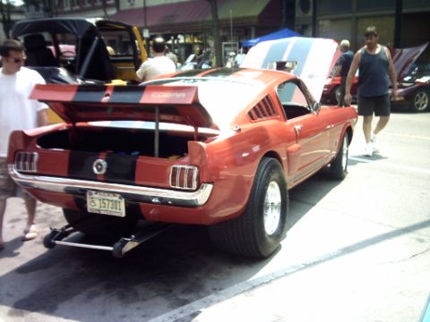 1995 Ford Probe - Red 1965 Ford Mustang fastback with black racing stripes and open trunk at an outdoor car show.