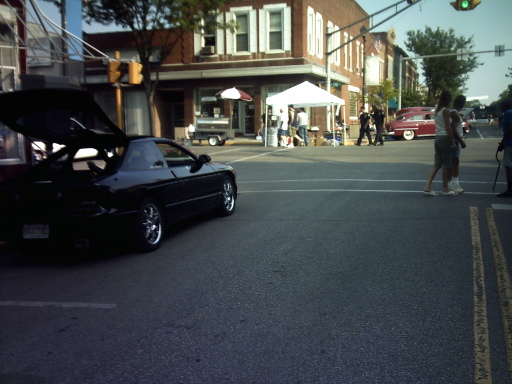 black coupe with trunk open, parked on street with people and tents nearby. Red classic car in background