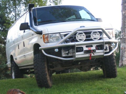 2000 Ford E350 - White Ford E-series van photographed from a 3/4 front angle, lifted on large mud-terrain tires with a tubular steel front brush guard bumpe…