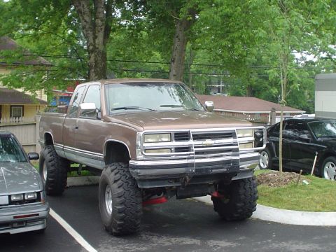 1981 Ford Bronco - Brown Chevrolet pickup truck with significant lift and oversized tires is parked in a lot.