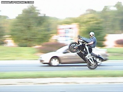 1994 Ford Bronco - A motorcycle is performing a wheelie alongside a car on a highway, with trees and buildings in the background.