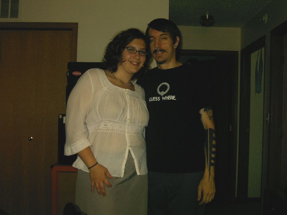Woman in white top and man in black t-shirt with 'GUESS WHERE.' text pose for photo. SUPERMOTORS.NET logo visible