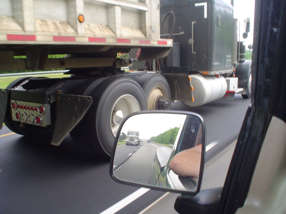 View from car window shows side of semi-truck trailer and reflection of another semi-truck in side mirror