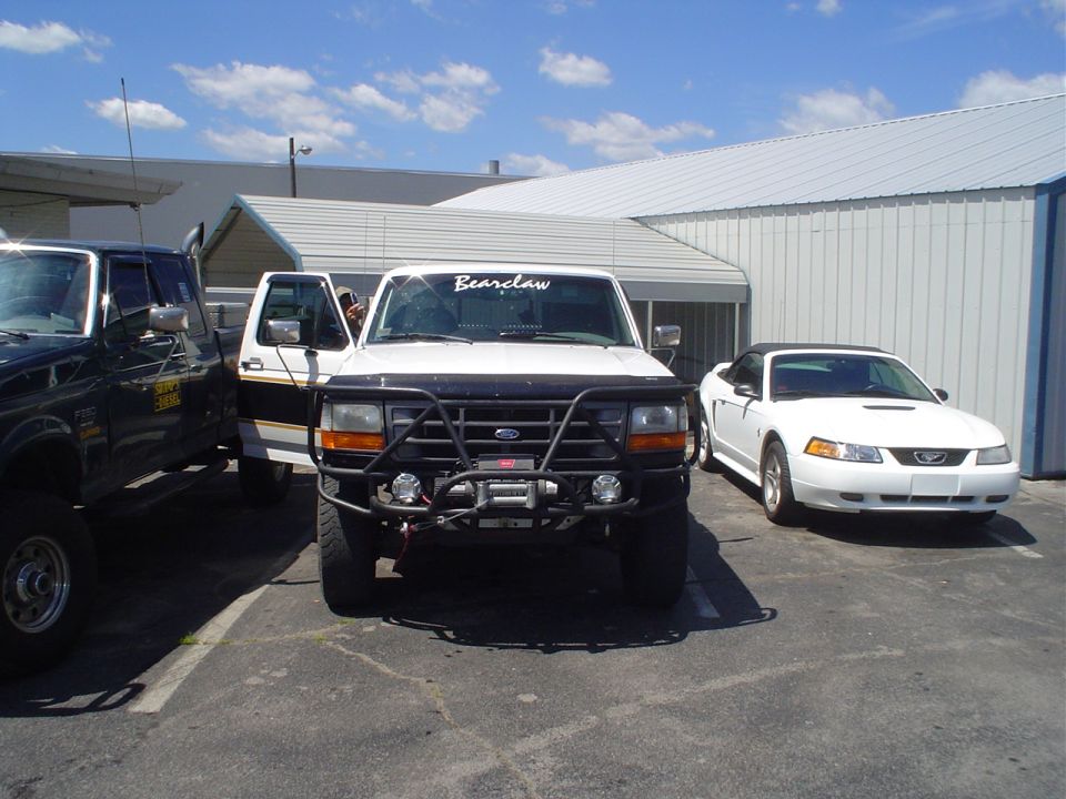 White Ford truck with bull bar and winch, parked next to a black truck and a white Ford Mustang convertible