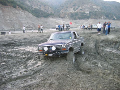 1985 Ford Bronco - A Ford Bronco navigates a muddy off-road course with other vehicles and spectators present.
